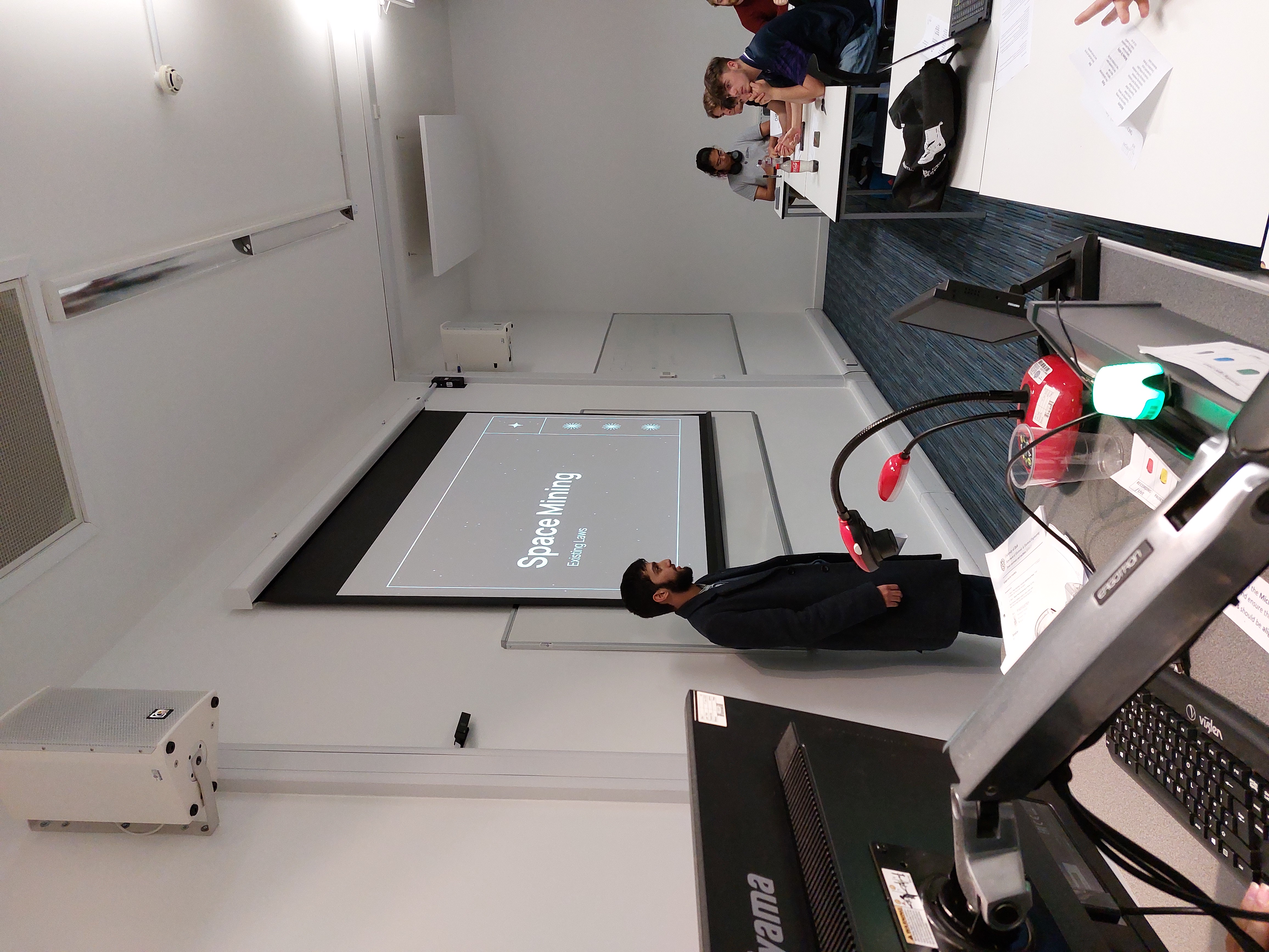 A student presents a slideshow titled 'Space Mining'. A relaxed audience watches with drinks and phones on their desks.