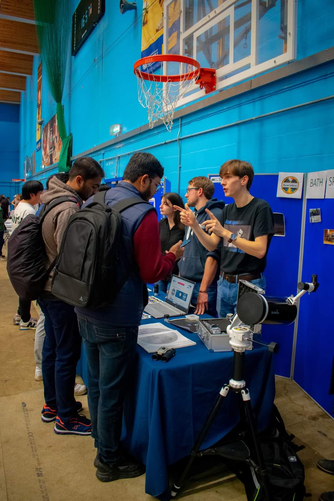 At the Groups Fair, the committee stands behind a table, decorated with telescope lenses, star charts and stickers. Two students chat to the committee from access the table.