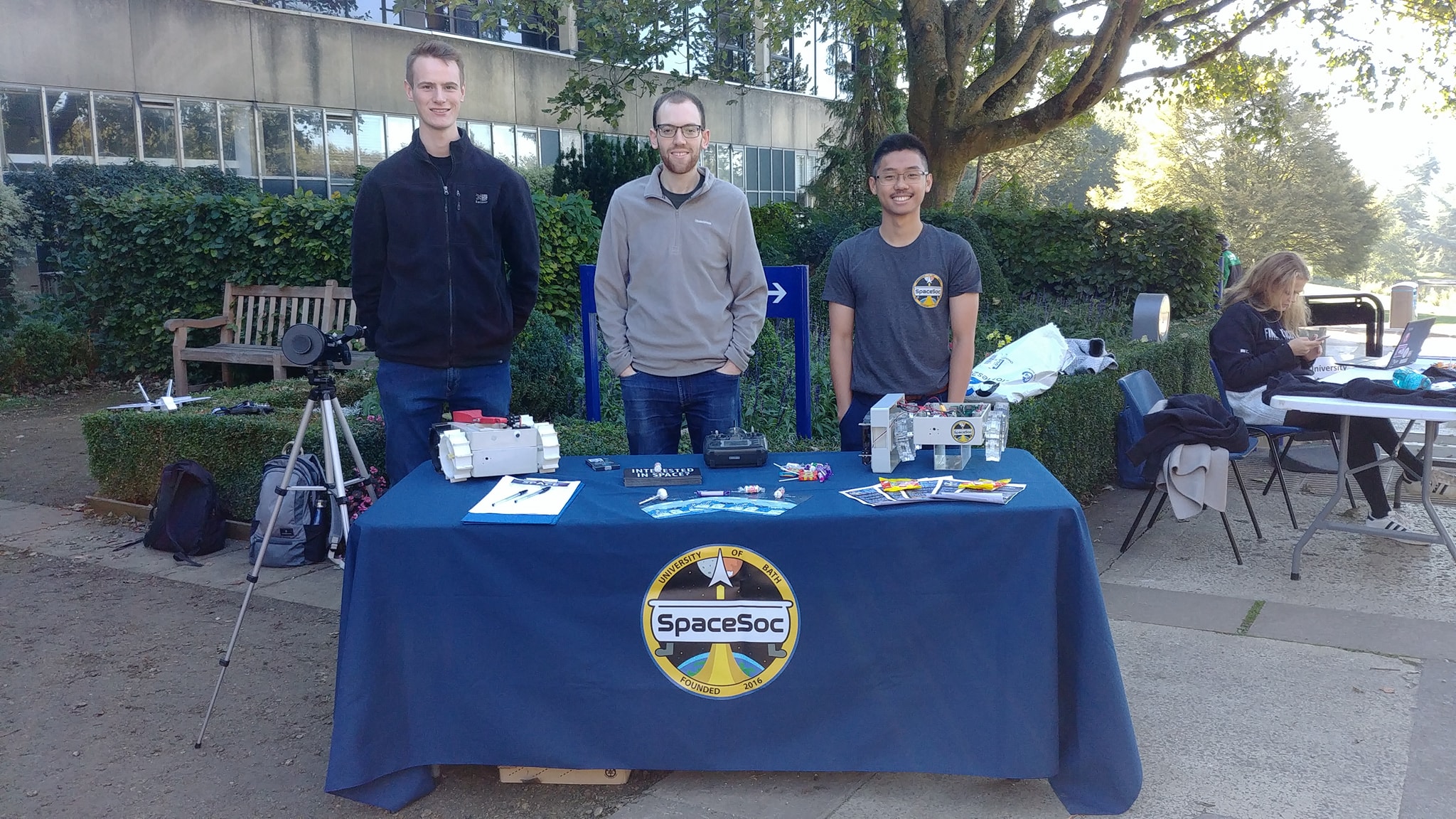 Three committee members stand behind a table for the 2018 Groups Day. Their table is decorated with rovers, flyers and sweets.