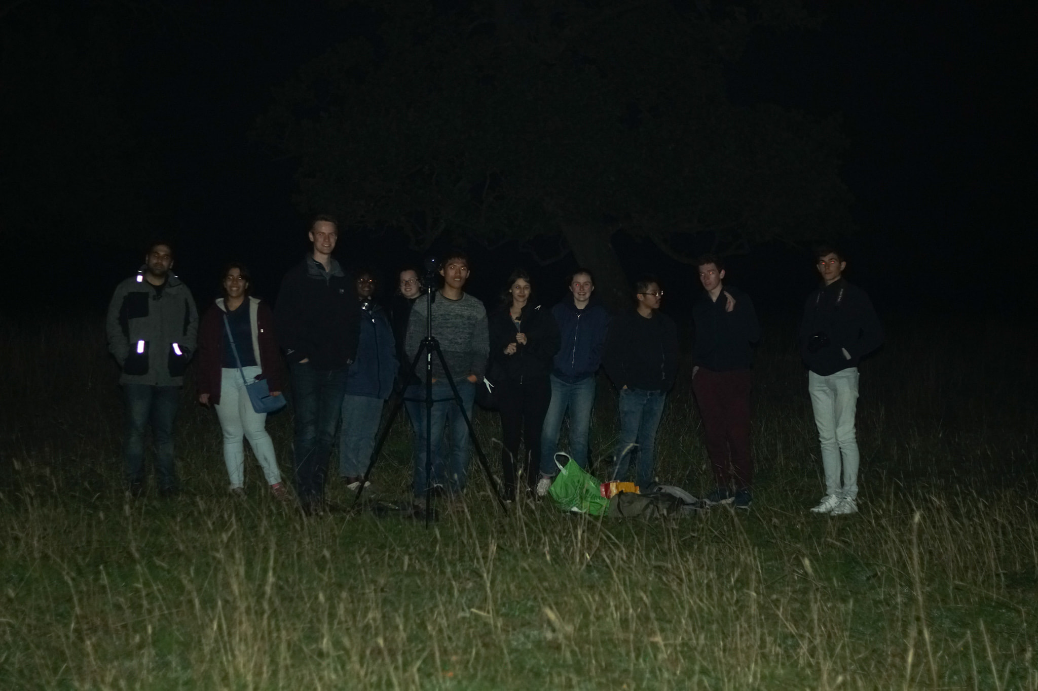 During a 2018 stargazing session, a dozen students stand in a row behind a small telescope. It is very dark.